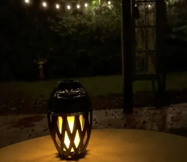 TikiTunes on a patio table at night with the LED flame glowing warm amber under string lights